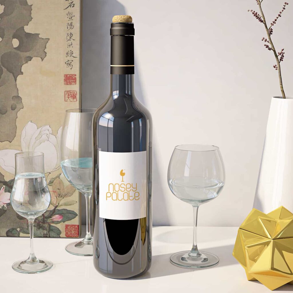 A bottle of wine with the new logo design and branding for Nosey Palate. The wine bottle is in the middle of 2 wine glasses on a white table.