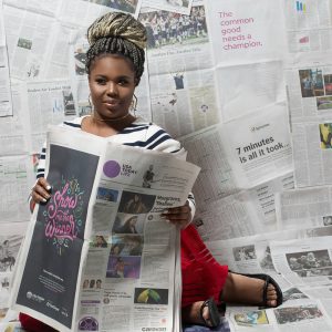 African American woman sitting down holding open newspaper with backdrop of newspapers